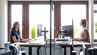 Zwei Frauen sitzen an Schreibtischen vor einem Fenster und arbeiten konzentriert an ihren Aufgaben.